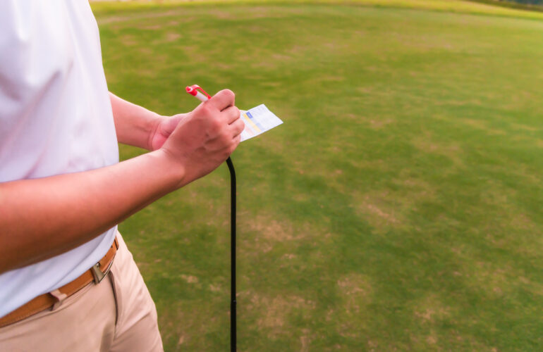 Asian male golfer using pen writing his golf score on scorecard while standing on green fairway.