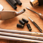 Professional golf club fitting tools including iron heads, shafts, ferrules, and a heat gun on a wooden workshop table.