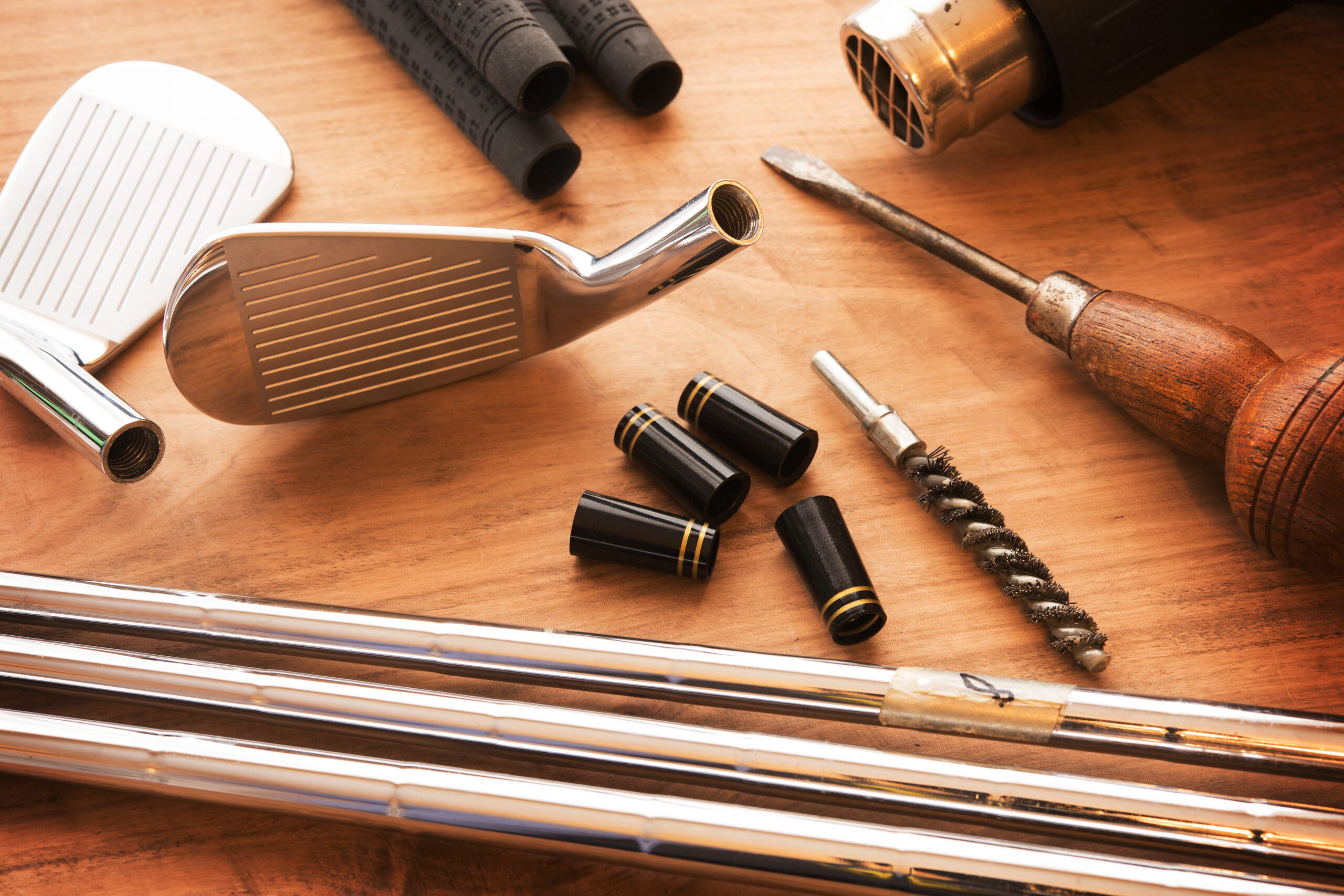 Professional golf club fitting tools including iron heads, shafts, ferrules, and a heat gun on a wooden workshop table.