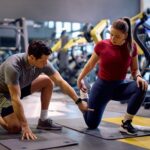 A personal trainer in a grey polo shirt is kneeling on a gym mat, guiding an athlete through a hip flexor stretch.