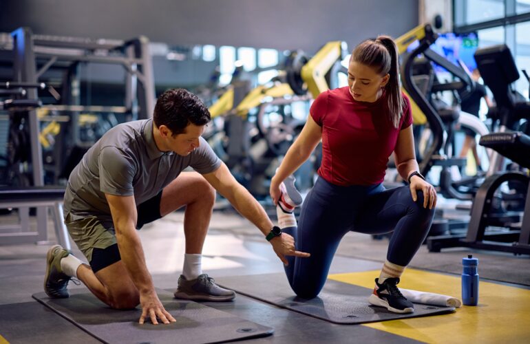 A personal trainer in a grey polo shirt is kneeling on a gym mat, guiding an athlete through a hip flexor stretch.