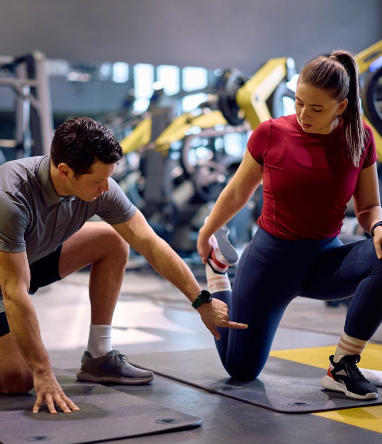 A personal trainer in a grey polo shirt is kneeling on a gym mat, guiding an athlete through a hip flexor stretch.
